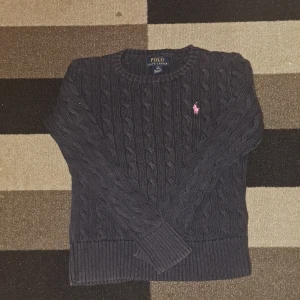 Ralph Lauren - Säljer en snygg mörkblå stickad tröja från Ralph Lauren med kabelstickat mönster och röd logga på bröstet. Tröjan har långa ärmar och ribbstickade kanter. Perfekt för kyliga dagar!