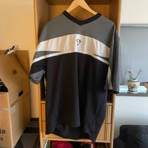 Y2k Svart och grå t-shirt med v-ringning - Snygg t-shirt i svart och grått med v-ringning och ett stilrent märke på bröstet. Den har korta ärmar och en bred vit rand över bröstet som ger en sportig look.