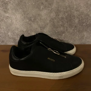 Svarta sneakers från Axel Arigato - Snygga svarta sneakers från Axel Arigato med dragkedja och rund metallring som detalj. Skorna har en vit sula och är tillverkade i skinn, vilket ger en stilren och modern look.