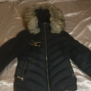Svart pufferjacka med päls från Hollies - Snygg svart pufferjacka från Hollies med avtagbar päls på huvan och leopardmönstrat foder. Jackan har dragkedja och flera praktiska fickor med gulddetaljer. Perfekt för kyliga dagar. Kan ha lite smink fläckar.