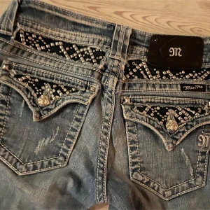 Jeans med strassdetaljer och slitningar - Snygga jeans från Miss Me med strassdetaljer på bakfickorna och slitningar längs benen. De har en klassisk blå färg och en lågmidjad passform. Perfekta för en trendig look.
