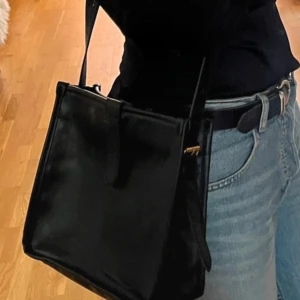 Svart axelväska i skinn - Snygg svart axelväska i skinn med enkel design. Perfekt för att bära dina dagliga nödvändigheter. Väskan har en justerbar axelrem och en praktisk dragkedja för säker förvaring.