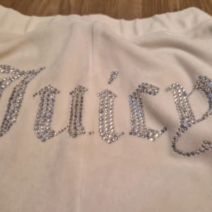 Vita mjukisbyxor med strassdetaljer från Juicy Couture - Säljer ett par vita mjukisbyxor från Juicy Couture med glittrande strassdetaljer. Byxorna har en avslappnad passform och är perfekta för en bekväm stil. Det ikoniska 'Juicy' är dekorerat på baksidan.