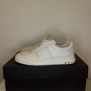 Vita Valentino flycrew - Snygga vita sneakers från Valentino med snörning och unika nitar på sulan. Skorna har en cool kamouflagemönstrad sula i grönt och svart. Perfekta för en stilren look.