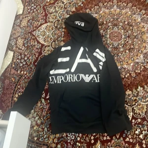 Svart hoodie från Emporio Armani - Snygg svart hoodie från Emporio Armani med stor vit logga på framsidan. Perfekt för en casual look med sin bekväma passform och huva. Ett måste för garderoben!
