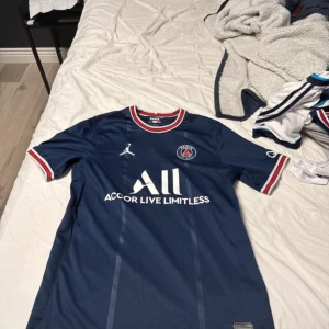 Blå PSG fotbollströja från Jordan - Snygg blå PSG fotbollströja från Jordan med korta ärmar och vita samt röda detaljer. Tröjan har klubbens emblem på bröstet och texten 'Accor Live Limitless' på framsidan. Perfekt för fotbollsfans! Äkta kosta 1100 ny pris kan diskuteras 
