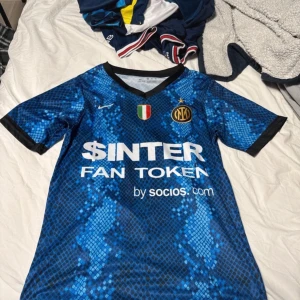 Blå fotbollströja från Nike - Säljer en blå och svart kortärmad fotbollströja från Nike med Inter-logga och sponsortryck. Tröjan har ett unikt mönster och spelarnamnet Džeko med nummer 9 på ryggen. Perfekt för fans av laget! Pris kan diskuteras