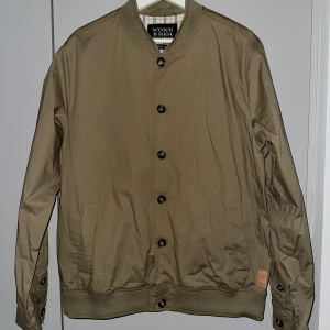 Beige/grön bomberjacka från Scotch & Soda - Snygg beige bomberjacka från Scotch & Soda med unikt blommönster på ryggen. Jackan har knappar framtill och ribbade muddar vid ärmslut och nederkant. 