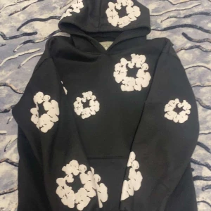Svart hoodie från Denim Tears - Säljer en svart hoodie från Denim Tears med vita blommönster. Den har en klassisk passform med huva och långa ärmar. Perfekt för en avslappnad stil.
