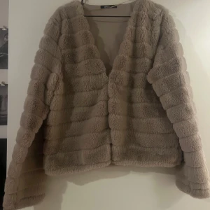 Beige pälsjacka från Gina Tricot - Mysig beige pälsjacka från Gina Tricot med långärmad design. Perfekt för kyliga dagar med sin fluffiga och mjuka textur. Jackan har en öppen front utan knappar eller dragkedja.