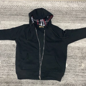 Svart huvtröja från Burberry - Säljer en stilren svart huvtröja från Burberry med dragkedja och klassiskt rutigt foder i huvan. Tröjan är tillverkad i bomull och har en bekväm passform med långa ärmar och praktiska fickor framtill. Storlek M men funkar för storlek S också priset in diskuteras 