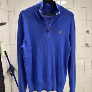 Blå half zip tröja från Ralph Lauren - Snygg blå tröja från Ralph Lauren med halv dragkedja och det klassiska broderade logotypen i orange på bröstet. Perfekt för en stilren look.