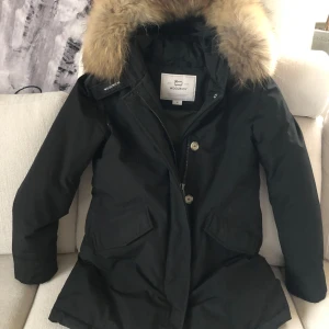 Svart parkajacka från Woolrich - Säljer en svart parkajacka från Woolrich med pälskantad huva. Jackan har dragkedja och knappar framtill samt praktiska fickor. Den är endast använd 1 vinter så i väldigt bra skick. 