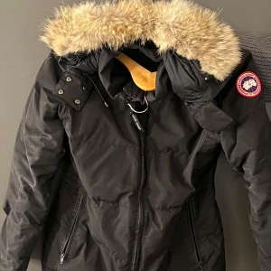 Svart dunjacka från Canada Goose - Säljer en svart dunjacka från Canada Goose med pälsfodrad huva. Jackan har en klassisk design med dragkedja och knappar framtill samt en broderad logotyp på ärmen. Perfekt för kalla vinterdagar.