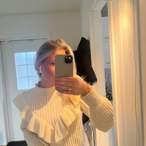 Beige stickad tröja med volangdetaljer - Säljer en snygg beige stickad tröja med volangdetaljer på framsidan. Tröjan har långa ärmar och en rund halsringning, perfekt för kyligare dagar. Passar bra till både jeans och kjol.