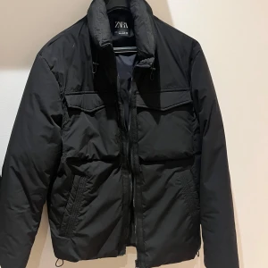 Svart pufferjacka från Zara - Snygg svart pufferjacka från Zara med dragkedja och hög krage. Jackan har långa ärmar och praktiska fickor framtill. Perfekt för kyligare dagar. Använd endast engång . 