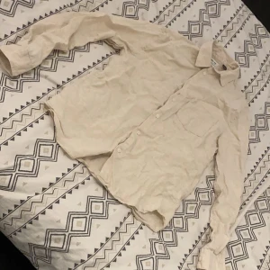 Beige linne skjorta - Säljer en stilren beige långärmad linnd skjorta med klassisk krage och knappar framtill. Perfekt för en avslappnad men ändå uppklädd sommar look. Skjortan har en bröstficka och är tillverkad i ett lätt material.