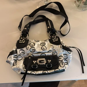 axelväska från Gussaci - As snygg axelväska från Gussaci med logotypmönster. Väskan har detaljer som dragkedjor och spännen samt en justerbar axelrem. Perfekt för att bära med sig det nödvändigaste med stil. köpt utomlands🫶