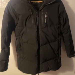 Svart dunjacka från Sail Racing - Säljer en svart dunjacka från Sail Racing med hög krage och dragkedja. Jackan har en logotyp på bröstet, armen & luvan samt flera praktiska fickor. Perfekt för kalla dagar med sin 500 fill power dunfyllning. Jackan var endast använd i 3 månader förra vintern. Köpt för 3.500 kr och säljer nu för 2.500 eftersom den är i ganska bra skick men ändå  använd. Anledningen till varför jag säljer den är för att jag vill köpa ny jacka, men annars var jackan jättebra och höll mig varm!