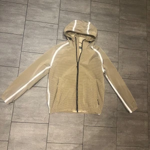 J Lindberg Windbreaker - J lindberg windbreaker i en snygg beige färg med vita detaljer. Perfekt inför våren då den är vattentät och står emot vind. Tveka inte på att lämna ett prisförslag! / Max 