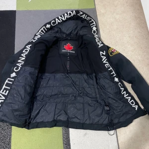 Svart pufferjacka från Zavetti Canada - Snygg svart pufferjacka från Zavetti Canada med broderad logga på bröstet och ärmen. Jackan har en huva med textdetaljer och dragkedja framtill. Perfekt för kyliga dagar.