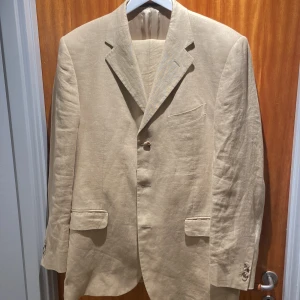 Beige linnekostym från Nino Danieli - Snygg beige kostym, kavaj och byxa i linne från Nino Danieli. Perfekt för varmare väder med sin lätta och luftiga känsla. Kavajen har klassiska slag och knappar framtill, samt fickor för en stilren look. 