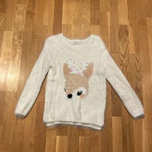 Vit fluffig tröja med renmotiv från H&M - Mysig vit tröja från H&M med ett gulligt renmotiv på framsidan. Renens horn är dekorerade med paljetter och pärlor, vilket ger en festlig touch. Tröjan är långärmad och har en fluffig textur som gör den perfekt för kyligare dagar. Också perfekt jul tröja ❤️