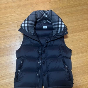 Burberry jacka - Snygg svart pufferjacka från Burberry med en rutig huva i grått och vitt. Jackan har dragkedja och knappar framtill samt praktiska fickor med dragkedjor. Perfekt för kyliga dagar.