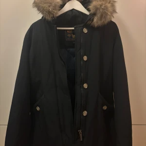 Svart vinterjacka från Woolrich - Säljer en svart parkajacka från Woolrich med pälskantad huva och knappar framtill. Jackan har en klassisk design med praktiska fickor och dragkedja. Perfekt för kyliga dagar.