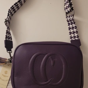  Helt ny Lila axelväska med mönstrad rem - Snygg lila axelväska från Bee & Dee med ett coolt mönster på remmen. Väskan har en dragkedja upptill och en extra ficka med dragkedja på baksidan. Perfekt för att ge din outfit en färgklick!