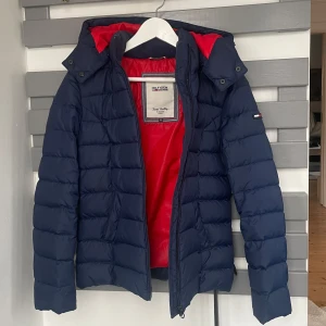 marinblå dunjacka från Tommy Hilfiger - mörkblå dunjacka från Tommy Hilfiger med röd insida och avtagbar huva. väldigt sparsamt använd, som ny! supervarm och lätt, 70% dun och 30% fjäder. storlek XS men passar även mig som vanligtvis har S. minns inte exakt vad den kostade men korta tommy hilfiger dunjackor brukar ligga runt 1700-2500kr!