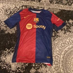 FC Barcelona fotbollströja - Snygg FC Barcelona tröja från Nike med korta ärmar. Tröjan har ett klassiskt röd- och blåmönster med sponsorloggor från Spotify och UNHCR. Perfekt för alla Barça-fans! 24/25