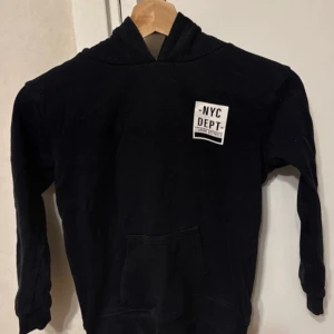 Svart hoodie med NYC-tryck - Säljer en svart hoodie med tryck 'NYC DEPT' på bröstet och 'BRNX/19' på ärmen. Den har en klassisk känguruficka och en bekväm huva. Perfekt för en avslappnad stil.
