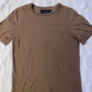 Brun t-shirt från Peak Performance - Säljer en brun t-shirt från Peak Performance med liten logga på bröstet. T-shirten är kortärmad och tillverkad i mjukt material, perfekt för en avslappnad stil.