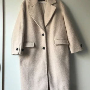 Beige kappa med knappar - Elegant beige kappa med långa ärmar och stora bruna knappar. Klassisk design med breda slag och fickor framtill. Perfekt för kyligare dagar. I ny Skick. Längd till småbenet.Oversize modell.