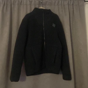 Svart fleecejacka från Under Armour - Säljer en svart fleecejacka från Under Armour med dragkedja framtill. Jackan har en hög krage och långa ärmar, perfekt för kyliga dagar. Den är stilren och bekväm, idealisk för både träning och vardag.