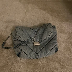 Grå quiltad axelväska med kedja - Snygg grå quiltad axelväska i jeansmaterial med kedjeaxelrem och metallspänne. Perfekt för en trendig look.