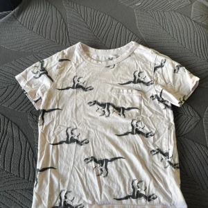 Vit t-shirt med dinosauriemönster från H&M - Säljer en cool vit t-shirt från H&M med ett svart dinosauriemönster. T-shirten har en bröstficka och korta ärmar. Perfekt för dig som gillar unika tryck!Använd på bild 4 fin en fläck på T-shirten