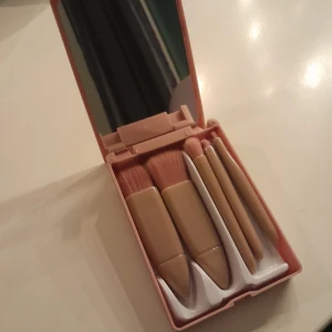 Sminkborstset i rosa etui - Praktiskt sminkborstset i ett kompakt rosa etui med inbyggd spegel. Innehåller fem borstar med beige handtag och mjuka borst, perfekt för olika sminkapplikationer.