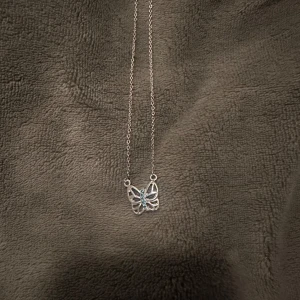 Silverfärgat halsband med fjärilshänge - Elegant silverfärgat halsband med ett detaljerat fjärilshänge. Hänget har vackra blåa sten i mitten som ger en fin kontrast. Perfekt för att lägga till en subtil touch av färg och elegans till din outfit.