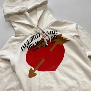 Vit hoodie från Helmut Lang med tryck - Säljer en vit hoodie sällsynt från Helmut Lang med ett stort rött hjärta och piltryck på framsidan. Hoodien har en klassisk design med dragsko i huvan och ribbade muddar vid ärmslut och nederkant. Säg till vid frågor!