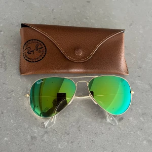 Ray-Ban Aviator Solglasögon med gröna spegelglas - Säljer ett par snygga Ray-Ban Aviator solglasögon med gröna spegelglas och guldfärgad metallram. De kommer med ett brunt fodral som har Ray-Ban-loggan på. Perfekta för soliga dagar och ger en cool look!
