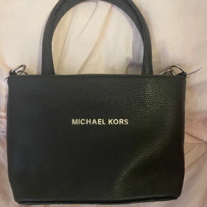 Svart handväska från Michael Kors - Säljer en snygg svart handväska från Michael Kors. Väskan är i läder och har en stilren design med silverfärgad logga på framsidan. Den har både handtag och en justerbar axelrem, vilket gör den superpraktisk. Insidan är fodrad med ett mönstrat tyg! 