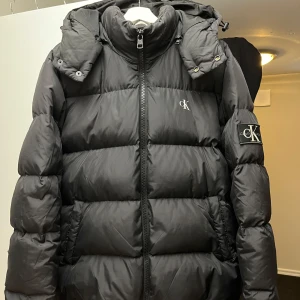 Clavin Klein vinterjacka herr - Snygg och skön jacka i använt skick. Huvan går att ta av med blixtlås om man vill det. Hål vid höger och vänster armbåge (se bilder), men går att laga/täcka över med någon lagnings lapp. 