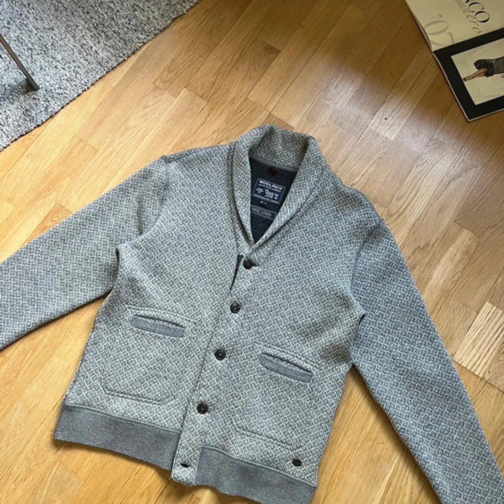 En grå cardigan från woolrich med mönster i grått och vitt. Den har en krage som går upp till halsen och knappar framtill. Den har två fickor på framsidan.   Knappt använd o perfekt nu till hösten. Tröjor & Koftor.