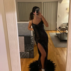 Svart glittrig klänning - En svart figursydd festklänning med paljetter på, en hög slits och fjädrar nedtill som släpar när man går, det är en klänning som passar perfekt till festliga tillfällen. Inköpspris: 2500