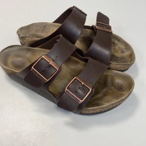 Bruna sandaler från Birkenstock - Säljer ett par klassiska bruna sandaler från Birkenstock med två justerbara remmar och kopparfärgade spännen. Sandalerna har en bekväm fotbädd och en slitstark sula. 