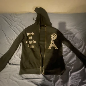 Svart hoodie med Eiffeltornet från Blinglucid - Snygg svart hoodie från Blinglucid med texten 'paris au clair de lune' och ett glittrande Eiffeltorn-tryck. Den har dragkedja och huva, perfekt för en cool och avslappnad stil. Knappt använd Max 5 gånger har bara legat och dammat typ om ni vill tvätta den så kolla in blinglucid hemsida så ni inte gör sönder diamanterna på tröjan