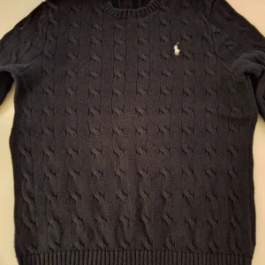 stickad tröja från Ralph Lauren - Säljer en stilren svart kabelstickad tröja från Ralph Lauren med deras ikoniska logga broderad på bröstet. Tröjan har en klassisk rund hals och långa ärmar, perfekt för kyligare dagar. Passar både till jeans och chinos för en avslappnad look.