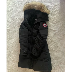 Svart parkajacka från Canada Goose - Säljer en svart parkajacka från Canada Goose med pälsdetalj på huvan. Jackan har en lång design med dragkedja och knappar framtill. Storlek S 